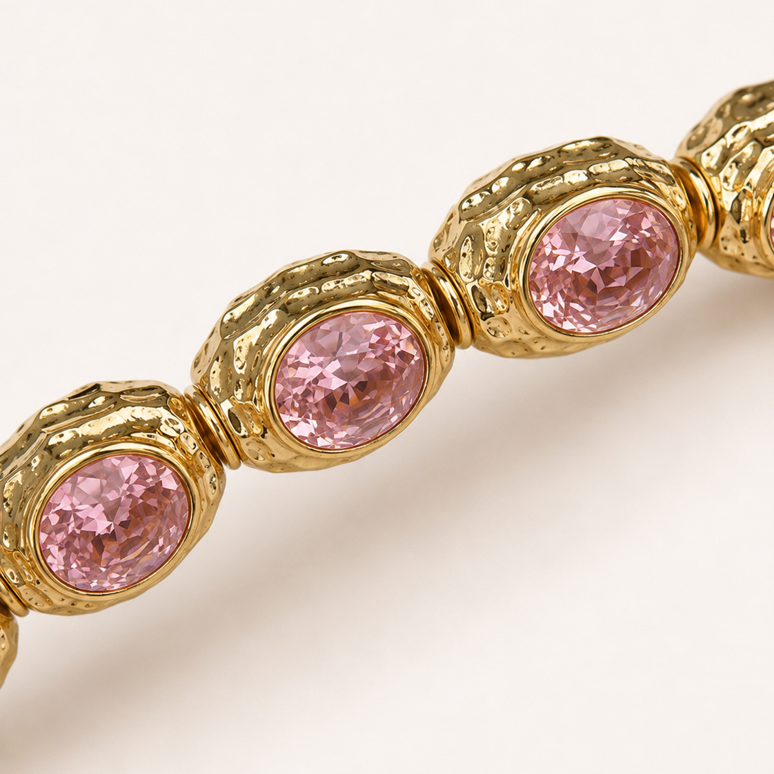 A close-up of a gold-toned bracelet featuring oval pink gemstones in textured bezel settings, shown against a clean off-white background.