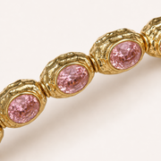 A close-up of a gold-toned bracelet featuring oval pink gemstones in textured bezel settings, shown against a clean off-white background.