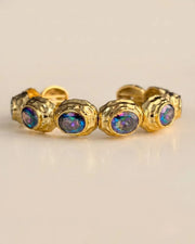 A gold-toned stretch band ring featuring five round iridescent opal-like gemstones, displayed on a clean, light beige background.