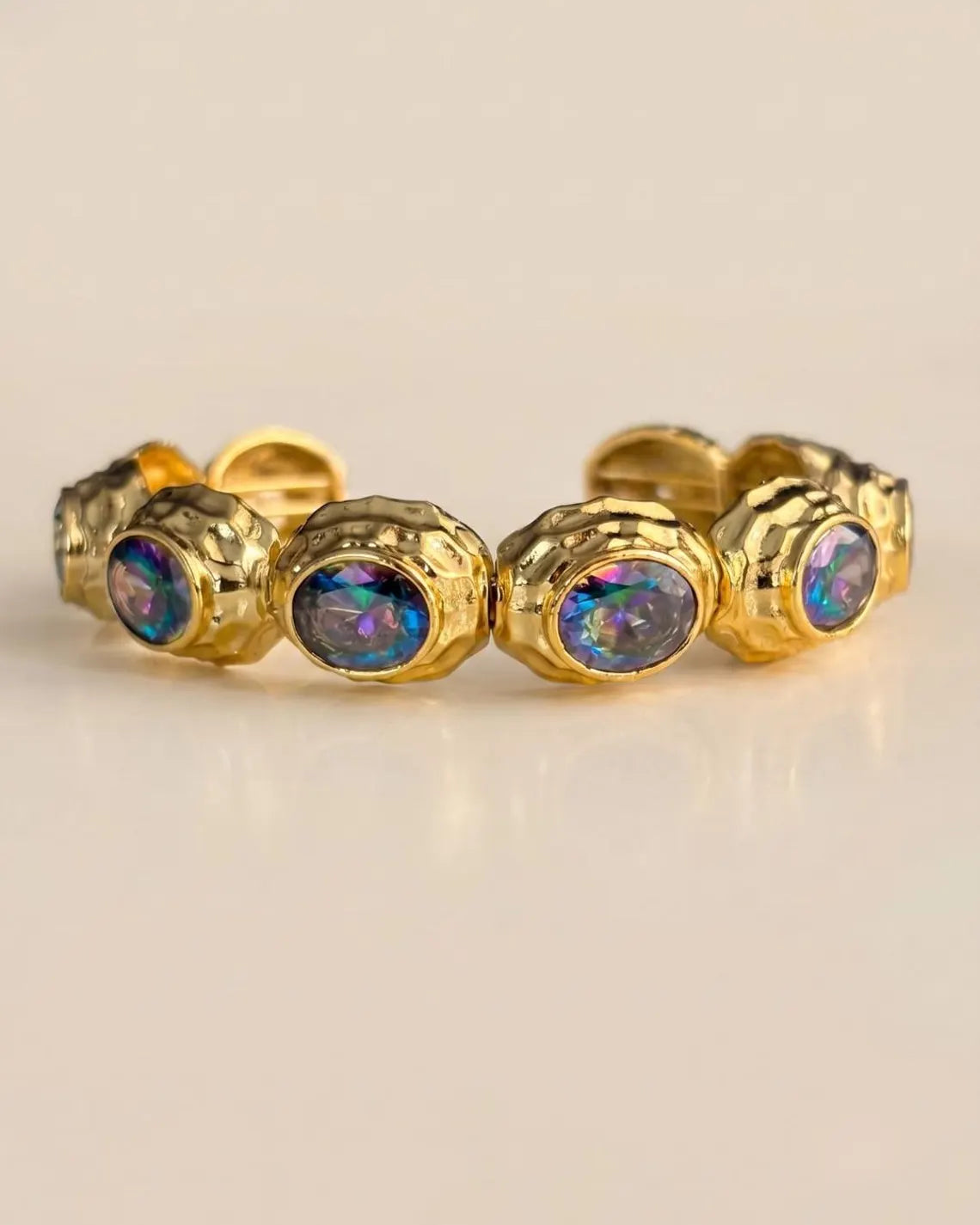 A gold-toned stretch band ring featuring five round iridescent opal-like gemstones, displayed on a clean, light beige background.