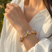 A gold-toned bracelet with oval light purple gemstones worn on a woman's wrist, shown in warm natural light against a white blouse.