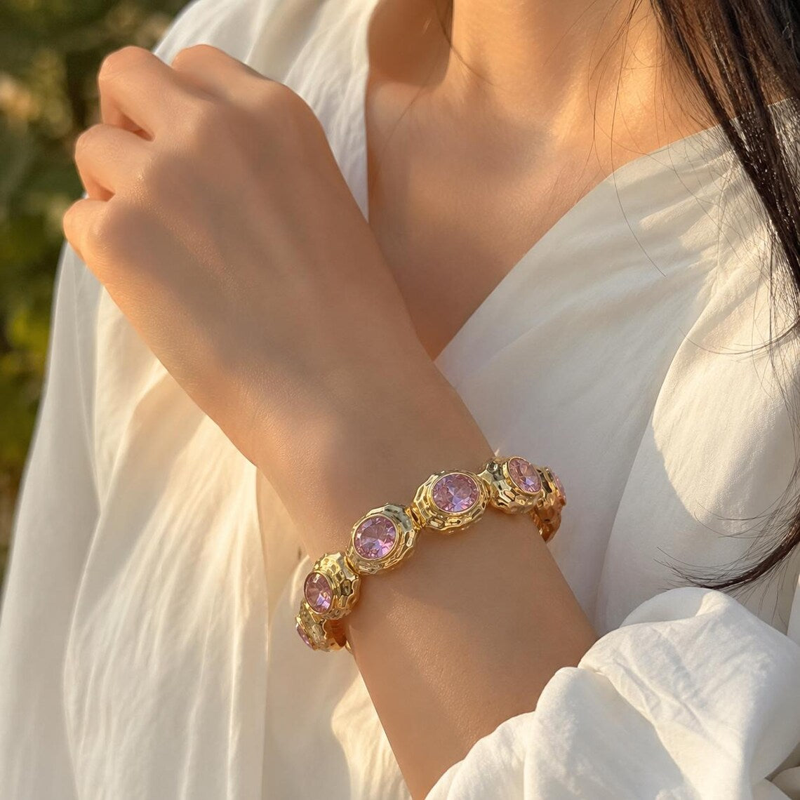 A gold-toned bracelet with oval light purple gemstones worn on a woman's wrist, shown in warm natural light against a white blouse.